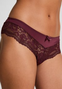 Nahaufnahme einer Person, die burgunderfarbene Spitzenbikini-Unterwäsche mit einer kleinen dekorativen Schleife am Bund trägt, vor einem neutralen Hintergrund.