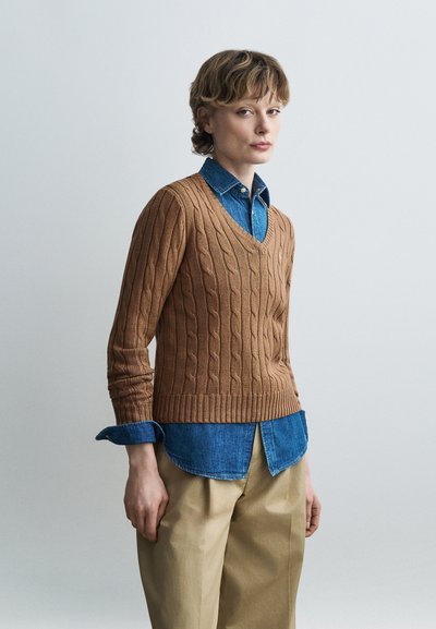 Personne portant un pull marron à col en V en maille torsadée sur une chemise en denim bleu avec un pantalon beige, debout devant un fond clair uni.