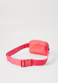 Ceinture de sac en tissu rose avec fermeture éclair, bandoulière réglable, et logo "HUGO" imprimé sur la bandoulière. Texture lisse, design compact.