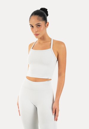 Kvinde med mørkt hår sat op i knold iført en hvid tætsiddende crop top med tynde stropper og matchende højtsiddende leggings, stående foran en ensfarvet baggrund.