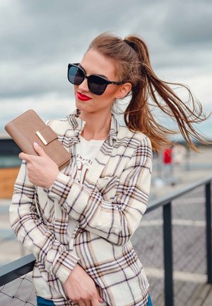 Beige and brown plaid shirt over a white T-shirt, holding a light brown wallet. Black oversized sunglasses. Long, wavy hair in a ponytail.
