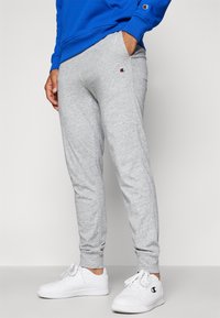Gray sweatpants made of soft fabric, featuring a cuffed ankle design and a small embroidered logo on the left thigh. Worn with white sneakers.