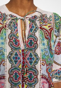 Multikolorová tunika s propracovanými paisley vzory v červené, zelené a modré barvě. Obsahuje výstřih ve tvaru klíče s dekorativními třásněmi.