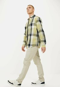 Skjorte med tern i gul, sort og hvid; har to brystlommer, knaplukning og lange ærmer. Sammen med lysebeige bukser og sneakers.