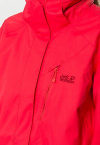Veste imperméable rouge avec un col montant, une poche avant zippée et un détail logo. Le matériau semble lisse avec une texture légère.