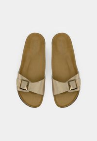 Ζεύγος μπεζ παντόφλες με πλατιά λουράκια και χρυσά αγκράφια, ταμπά πέλματα και μαύρες σόλες, φαίνονται από πάνω σε λευκό φόντο.