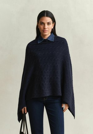 Frau mit langen dunklen Haaren trägt einen dunkelblauen Kabelstrick-Poncho über einem Jeanshemd und dunklen Jeans, hält schwarze Träger in der linken Hand.