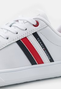 Bílá syntetická teniska s červenými a námořnickými pruhy, s označením značky a bílými tkaničkami. Texturovaná gumová podrážka a detaily s průchodkami.