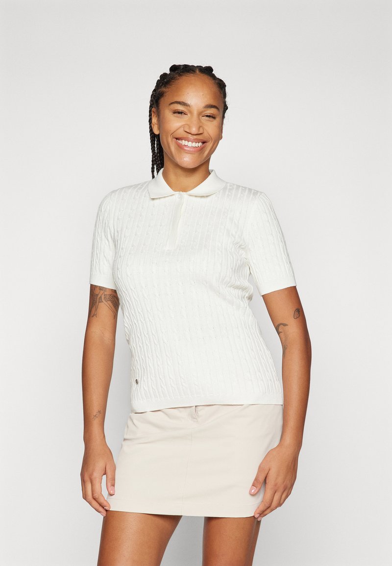 White short-sleeve polo with textured cable knit pattern and a collared neckline. Worn with a beige skirt, featuring a relaxed fit.