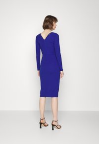 Vestido de manga longa, até ao joelho, em azul royal sólido com costas em V e detalhes em renda. Silhueta ajustada combinada com saltos abertos pretos.