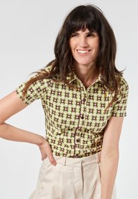 Camicia a maniche corte verde con stampa geometrica in viola e bianco. Presenta una chiusura con bottoni e un colletto. Tessuto dal tocco morbido.