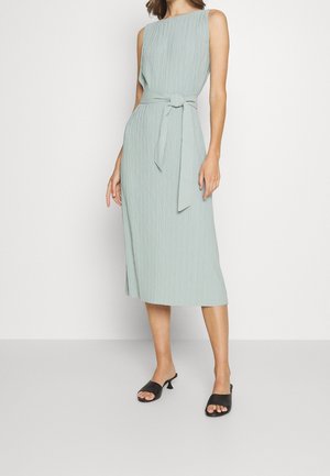 Bezruké světle zelené texturované midi šaty s látkovým páskem uvázaným v pase, nošené s černými otevřenými nízkými nazouváky na neutrálním pozadí.