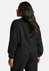 Svart, kortärmad topp med långa ärmar, tillverkad av mjukt material; har en avslappnad passform, rund halsringning och elastiska ärmslut.