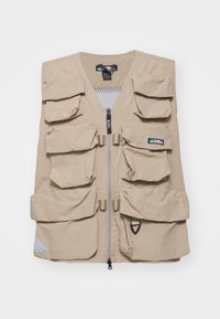 Beige Utility-Weste mit mehreren Vorderaschen, einem Reißverschluss, einem Netzfutter und einem kleinen Logoetikett auf der oberen linken Seite.