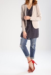 Beżowa marynarka narzucona na granatową sukienkę w kropki, zestawiona z przetartymi niebieskimi dżinsami i czerwonymi sandałami na wysokim obcasie. Gładkie materiały i dopasowany krój.