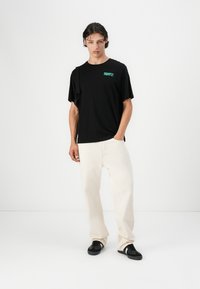 Camiseta negra de manga corta con el logo verde "ROOTS", combinada con pantalones de pierna ancha de color crema y zapatillas negras. Atuendo simple y casual.