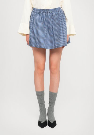 Jupe à carreaux bleus et blancs avec une taille élastique, au design superposé, associée à des chaussettes grises à hauteur de genou et des chaussures noires à bout pointu.