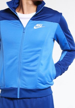 Person trägt eine blaue Nike-Sportjacke mit Reißverschluss und einem weißen Nike-Logo auf der Brust sowie passende blaue Hosen, Hände in den Taschen.