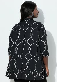 Donna con lunghi capelli neri indossa una tunica nera con linee bianche ondulate, grandi orecchini a cerchio in argento, in piedi di profilo contro una parete bianca.