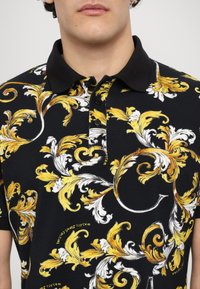 Polo noir avec un motif floral audacieux jaune et blanc, doté d'un col classique et d'une patte de boutonnage à trois boutons.