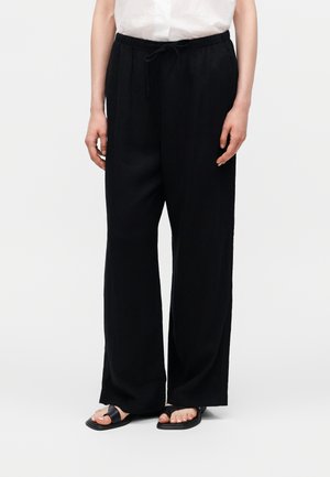 Osoba nosząca luźne czarne spodnie z ściągaczem w talii, biały top i czarne płaskie sandały na jednolitym jasnym tle.