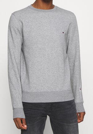 Mann trägt hellgraues Sweatshirt mit Rundhalsausschnitt und kleinem Logo auf der Brust und am Ärmel, kombiniert mit dunkelgrauen Jeans.