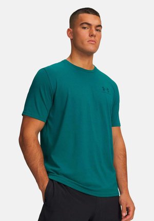 Uomo con capelli corti che indossa una maglietta a maniche corte Under Armour turchese e pantaloni neri, in piedi con una mano in tasca su sfondo bianco.