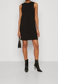 Sort ærmeløs kjole med tætsiddende pasform, der har en rund halsudskæring og kædedetalje. Matchet med sorte spidse ankelstøvler og en lille taske.