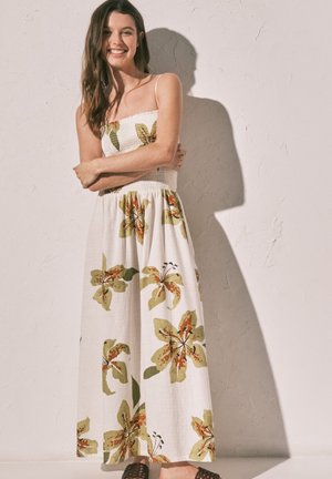 Usměvavá žena s dlouhými vlasy v bílé maxi šatech bez ramínek s velkými zelenými a oranžovými květinovými vzory stojící před strukturovanou zdí.