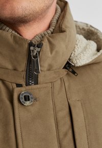 Braune Jacke mit Reißverschluss und hohem Kragen, mit Fleece-Futter und strukturiertem Außenmaterial. Verfügt über ein rundes Logo und sichtbare Nähdetails.