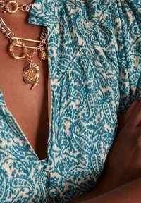 Camicia patterned in tessuto blu e bianco con collo a camicia, caratterizzata da dettagli con bottoni e sottili accenti metallici. Collana a catena d'argento spessa con charm dorati.