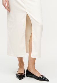 Πόδια που φορούν ένα λευκό midi φούστα με κεντρική σχισμή και μαύρες μυτερές φλατ με μικρά φιόγκους, στέκονται σε λευκό πάτωμα.