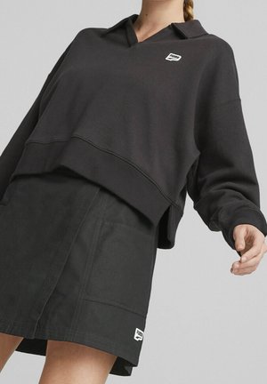 Person iført sort Puma sweatshirt med krave og matchende sort nederdel, stående mod en ensfarvet grå baggrund.