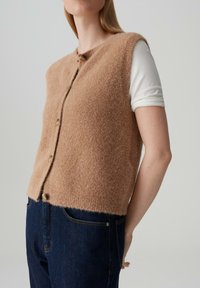Maglione senza maniche di maglia color beige, morbido e fluffy, con collo rotondo e bottoni frontali, abbinato a jeans blu scuro. Finitura morbida e testurizzata.