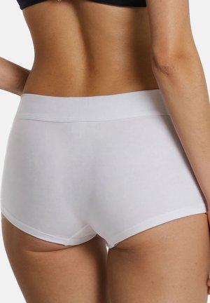 Kobieta ubrana w białe bezszwowe, wysokie bokserki, widziana od tyłu na jednolitym tle.