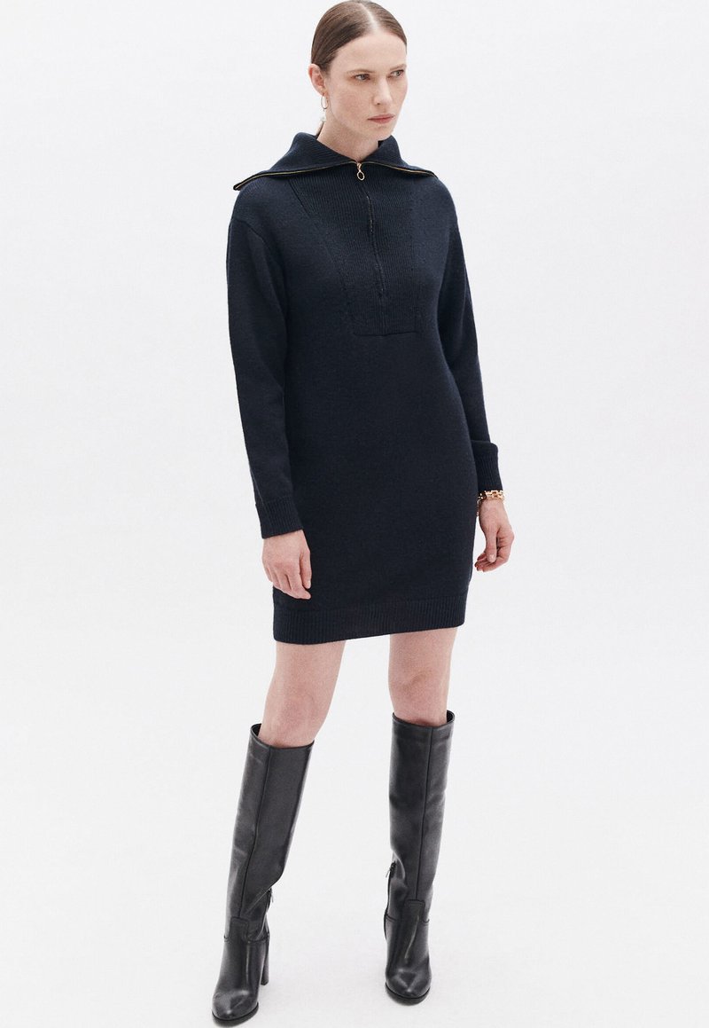 Vestito lavorato a maglia blu navy con collo alto e chiusura con zip; presenta maniche lunghe e orlo a coste. Abbinato a stivali neri al ginocchio.