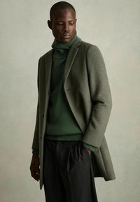 Pulover din lână de culoare verde închis, cu guler înalt, purtat sub o haină din lână de culoare măslinie deschisă, combinat cu pantaloni negri cu pliuri. Textură a materialului neted.