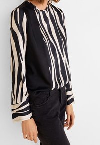 Frau, die eine langärmelige Bluse mit schwarz-beigem Zebra-Streifen trägt, kombiniert mit schwarzen hochtaillierten Jeans vor einem schlichten Hintergrund.