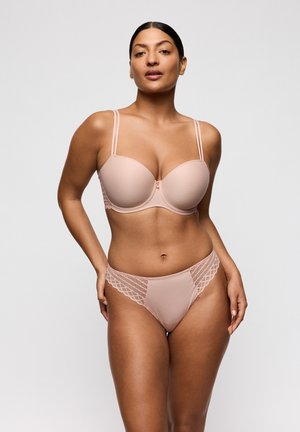 Vrouw met donker haar, een huidkleurige beha met dubbele bandjes en bijpassende slip met kantaccenten, staand tegen een effen lichte achtergrond.