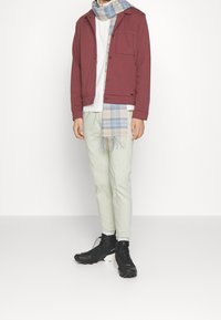 Mörkröd jacka, vit skjorta, ljusgrön avsmalnande byxor och svarta kängor; med en blå och beige rutiga scarf med fransar.