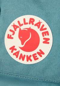 Fjällräven Kanken oznaka na tirkiznom materijalu. Okrugla oznaka prikazuje siluetu crvene lisice s tekstom "Fjällräven Kanken" u crvenoj boji oko nje.