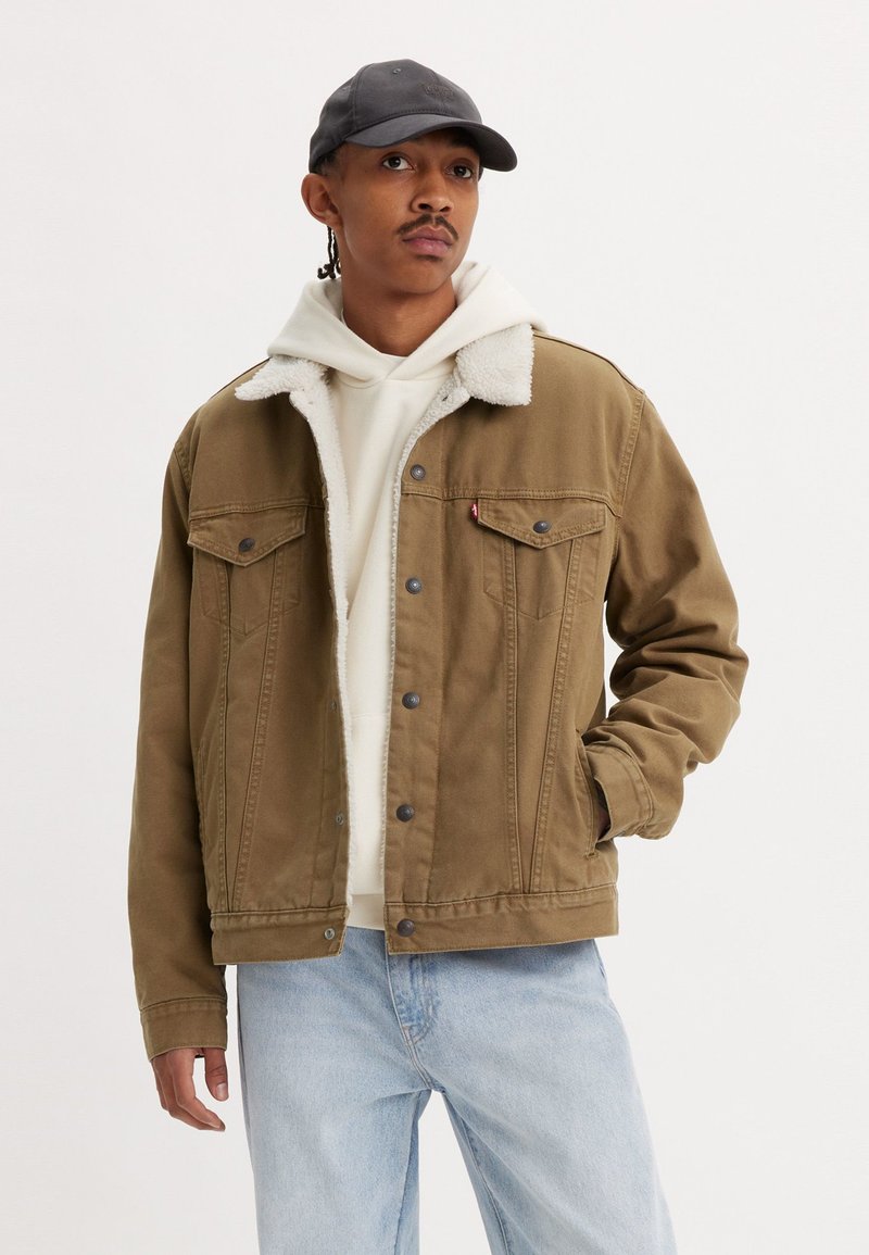 Veste en denim marron avec un col en fausse fourrure blanche, devant à boutons, deux poches poitrine, associée à un sweat à capuche blanc et un jean bleu clair.