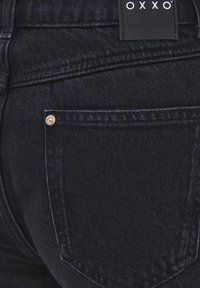 Vaqueros de mezclilla negra con un bolsillo trasero que incluye un detalle de remache metálico y una etiqueta con el nombre de la marca "OXXO". Textura suave, diseño ajustado.