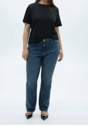 Personne portant une chemise noire à manches courtes, un jean bleu et des chaussures noires à bout pointu, debout devant un fond blanc.