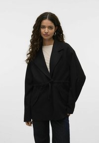 Must värvi suur ülerõivaste mantel pehmest kangast, millel on sidumisvöö, sälguga krae ja ees olevad taskud. Kanti heledat värvi pluusi peal.