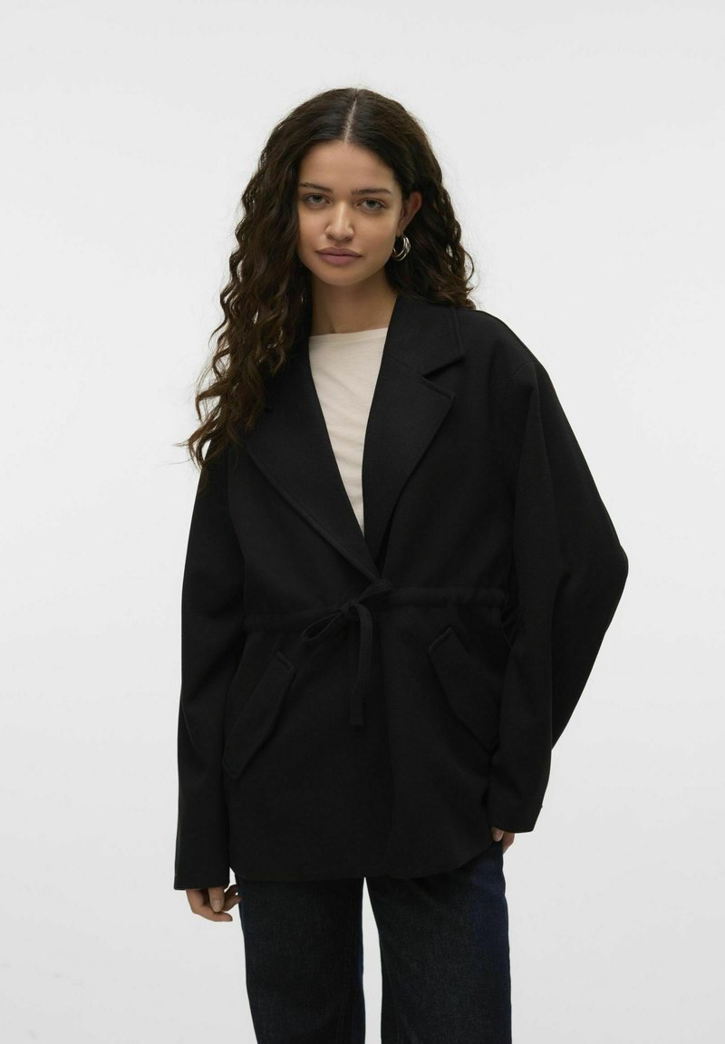 Must värvi suur ülerõivaste mantel pehmest kangast, millel on sidumisvöö, sälguga krae ja ees olevad taskud. Kanti heledat värvi pluusi peal.