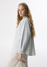 Camicia di jeans azzurro chiaro con maniche lunghe, tasca frontale e borchie decorative argentate sulle spalle. Presenta una vestibilità rilassata e un orlo curvo.