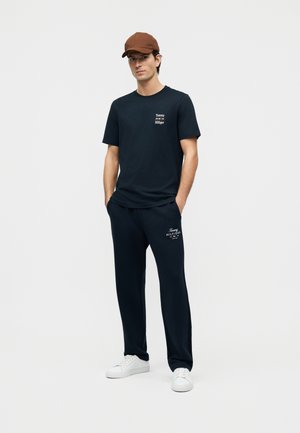 Man wearing a brown cap, black Tommy Hilfiger T-shirt and pants, both with white logo, and white sneakers, standing with hands in pockets.