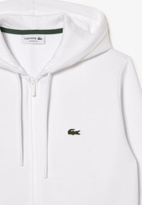 Sweat à capuche blanc zippé avec cordons et logo crocodile Lacoste vert sur la poitrine gauche, étiquette coupe classique à l'intérieur du col.