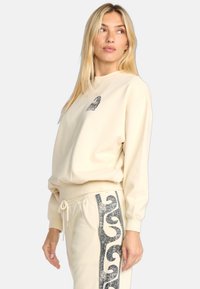 Felpa beige con scollo rotondo e logo sul petto, abbinata a pantaloni della tuta coordinati con un motivo scintillante scuro sulla parte laterale.